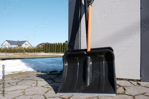 Two snow shovels leaning against a garage with melting snow, ready for winter cleanup. Residential scene highlighting snow removal tools and seasonal yard maintenance in early spring under blue sky.