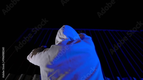Nighttime protest. Protester in white hoodie grips fence, shouts. Back view, low angle. Urban streets, dramatic tension, cinematic motion. Anonymous activist, moody atmosphere.