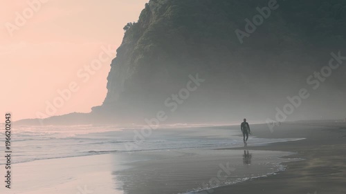 A surfer walks along the beach with his board at Karekare Beach in Auckland, New Zealand. He is walking toward the ocean to surf the waves at sunset.