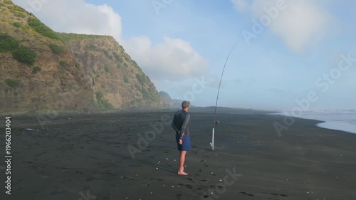 Auckland, New Zealand: A lone fisherman stands on the black sands of Karekare Beach, casting his line into the ocean, hoping for a catch on a misty morning.
