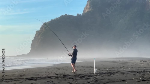 Auckland, New Zealand: A man is surfcasting on a beach with black sand. He is fishing for recreation and to enjoy the outdoors at Karekare Beach.