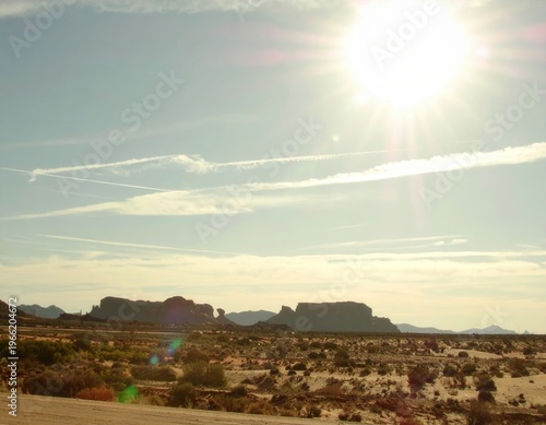 Paesaggio desertico arido sotto un sole splendente con formazioni rocciose e scie di condensazione nel cielo azzurro.