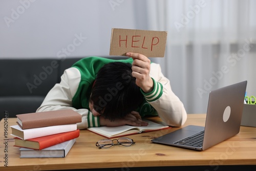Tired student holding cardboard sign with word Help before exam at wooden desk indoors