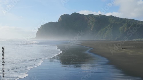Scenic view of people walking on the black sands of Karekare Beach, New Zealand, with the iconic Lion Rock in the background, creating a serene coastal landscape.