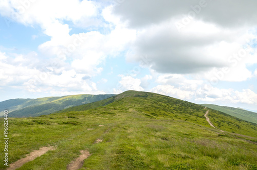 Sunlit green mountain ridge with a winding dirt trail leading over rolling hills under dramatic summer clouds. Svydovets, Carpathian Mountains, Ukraine