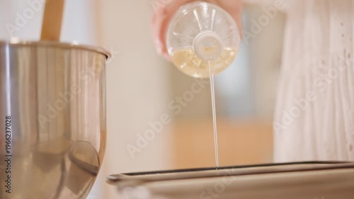Hands drizzling oil into pan before filling, clear plastic bottle releasing thin golden stream, mixing bowl and wooden spoon in background, careful measured pour, warm kitchen prep for baking