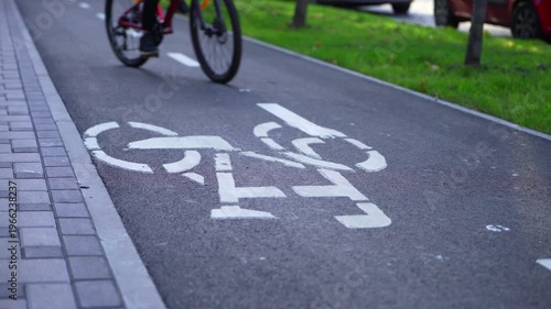 Detail of bicycle wheels moving along a marked bike lane in city park. Traffic symbol visible on asphalt. Slightly unsteady motion suggests beginner rider. Urban cycling safety concept.