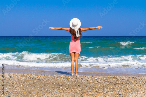 Back view stressfree carefree woman in pink dress, white hat standing barefoot on the beach, breathing fresh air enjoying summer. Travel, vacation, freedom, relaxation concepts.