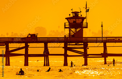San Clemente Pier at sunset with surfers and paddleboarders silhouetted in golden Pacific Ocean, Southern California