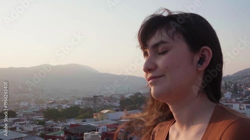 Beautiful young latin woman enjoying the wind on a rooftop, closing her eyes while listening to music with her earbuds at sunset over a city in mexico