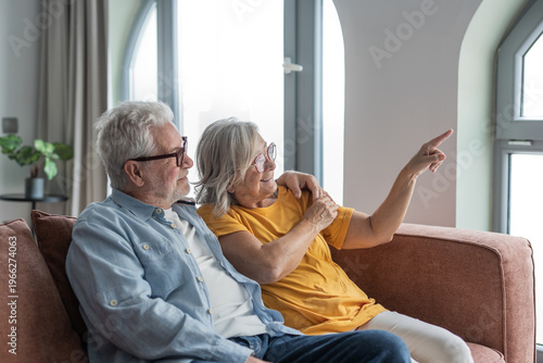 Tableau sur toile Senior couple relax on a couch, smiling as they point toward a bright window and share a tender moment of companionship, affection and contentment in their cozy living room