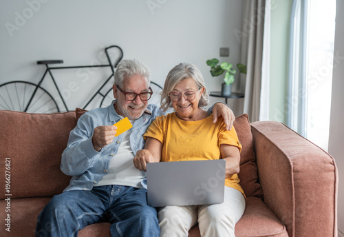 Obraz na plátně Happy senior couple sits on a comfortable sofa using a laptop and credit card to shop online, sharing a warm moment of connection, convenience and confidence in digital payments at home
