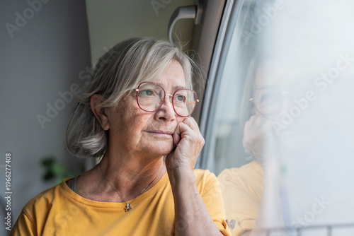 An elderly woman with grey hair and glasses gazes out the window, resting her chin on her hand. Soft natural light highlights her thoughtful expression and quiet reflective mood at home.
