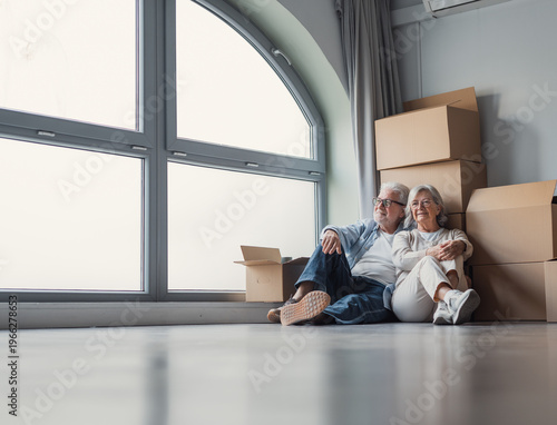 Fotografie A content senior couple sits on the floor surrounded by cardboard moving boxes in their bright new home, embracing a fresh start together and sharing a quiet, hopeful moment