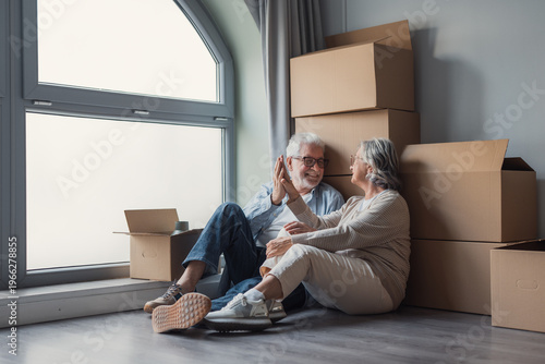 Foto Senior couple relax and celebrate moving day while seated on the floor surrounded by packed boxes, sharing a warm high-five as they begin unpacking and settle into their new home