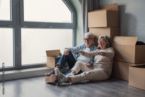 Фотография A happy senior couple relaxing on the floor surrounded by moving boxes in their new home, smiling and imagining furniture placement while taking a break during the relocation process
