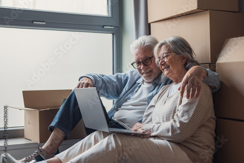 Obraz na płótnie An affectionate senior couple sits on the floor among moving boxes, smiling and using a laptop as they unpack and settle into their new home, sharing a relaxed, joyful moment together