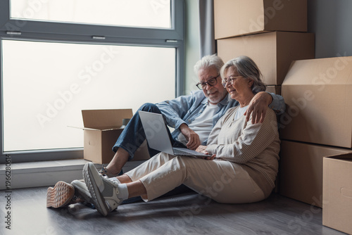 Obraz na płótnie Senior couple sits on the floor surrounded by packed boxes, using a laptop and smiling as they organize their move into a new home, sharing a quiet, happy moment and companionship