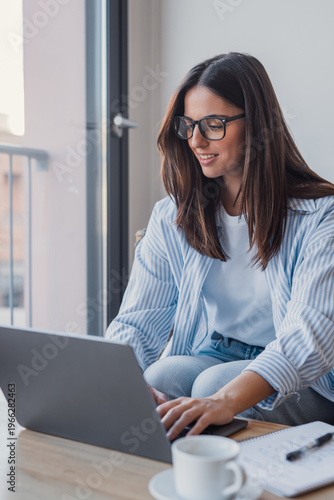 Young woman sits at a table by a window, smiling as she types on a laptop. Casual remote work setup with notebook, coffee cup and natural light conveying focus, comfort and productivity.