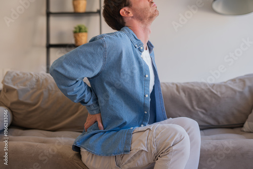 Young man sits on a sofa in a living room clutching his lower back in visible pain and discomfort, suggesting back strain, poor posture or sudden injury while at home. Casual clothing, daytime scene.