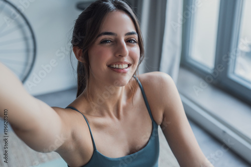 Smiling young woman takes a bright selfie in workout clothes by a large window, conveying casual fitness, confidence and healthy lifestyle at home. Natural light, relaxed mood and active wellbeing. Free Download Smiling young woman takes a bright selfie in workout clothes by a large window, conveying casual fitness, confidence and healthy lifestyle at home. Natural light, relaxed mood and active wellbeing. - GFXLoad