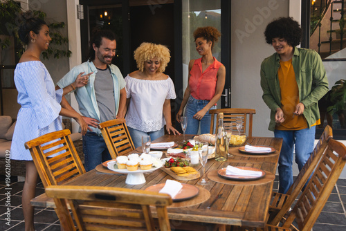 Diverse friends gathering around wooden dining table on covered patio, sharing cupcakes and wine