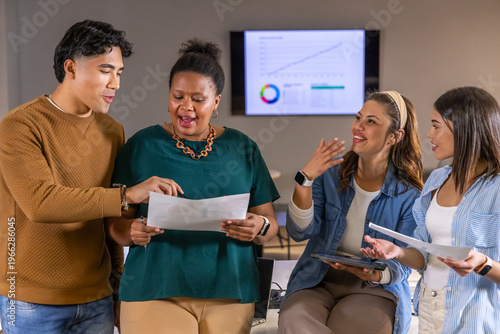 Diverse coworkers reviewing papers and tablet in office with wall display, in casual attire