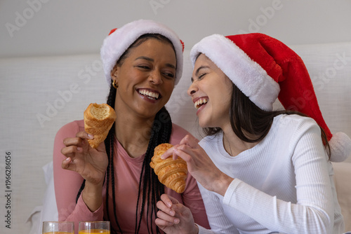 Diverse female friends laughing holding croissants wearing Santa hats on bed with orange juice