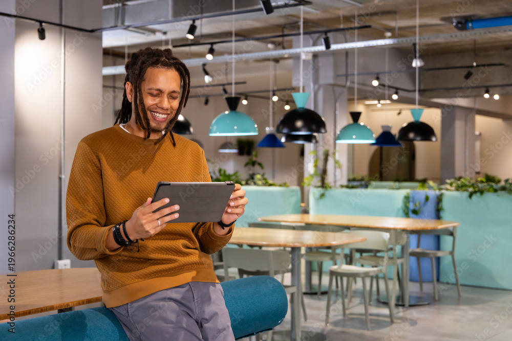 Fototapeta premium African American man in 30s wearing rust sweater leaning on teal bench using tablet in workspace