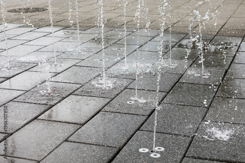 Foto Water jets shoot upward from the ground in a modern splash fountain, creating playful streams over gray stone tiles