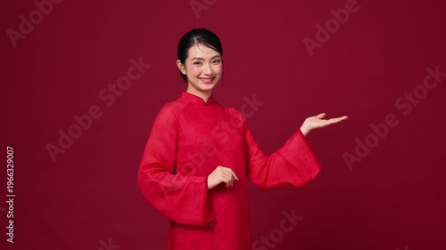 Asian woman pointing to the side, wearing red festive dress, ideal for product placement or holiday announcement design.