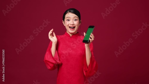 Asian woman celebrating with smartphone raised, joyful expression, red holiday background for digital promotion.