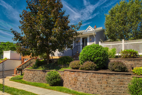 Bright exterior of a single-family home featuring a tiered masonry retaining wall, manicured shrubs, and mature trees. 