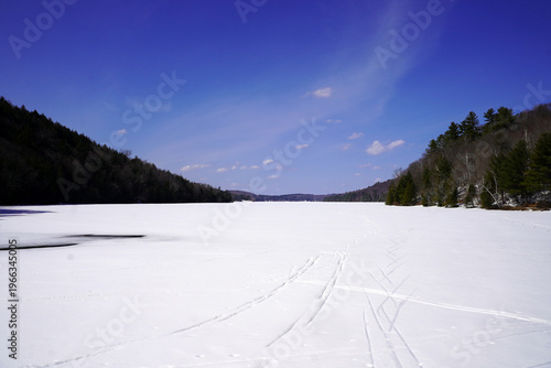 A vast, snow-covered expanse of frozen Lac Philippe is framed by densely forested hills under a vibrant blue sky in Gatineau Park, Quebec