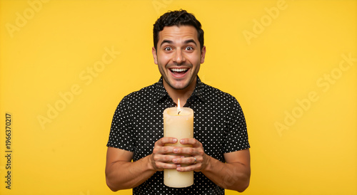 Surprised man holding a large lit pillar candle against a yellow background. Happy male in polka dot shirt with a burning candle. Celebration or inspiration concept