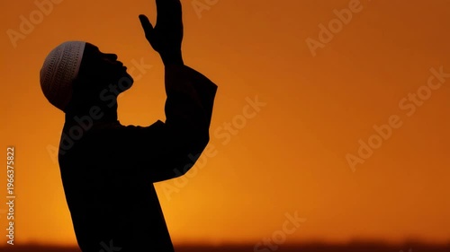 Silhouette of a person praying with raised hands against orange sky