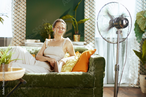 A middle-aged woman, eyes closed in serenity, relaxes on a green velvet couch with a book, enjoying the cooling breeze from a standing fan in her bright, plant-filled living room.