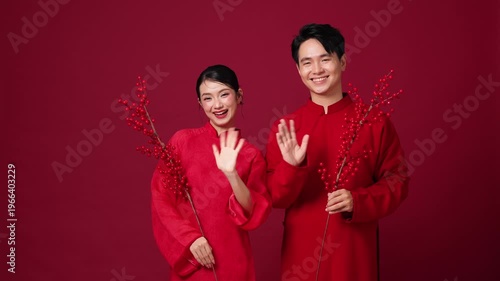 The Asian couple held branches of blooming flowers, waving their hands in greeting. Elegant red attire created a warm, festive atmosphere