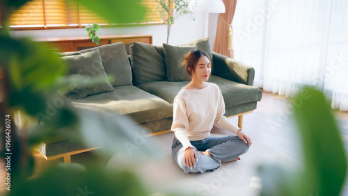Happy young Asian woman practicing yoga and meditation at home sitting on floor in living room in lotus position and relaxing with closed eyes. Mindful meditation and wellbeing concept