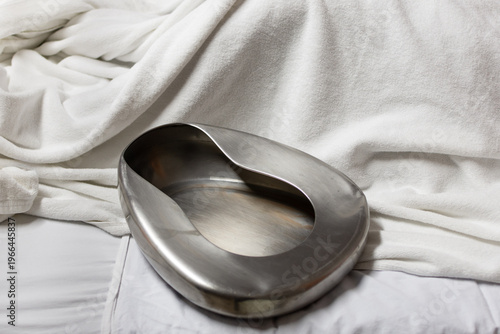 Stainless steel bedpan on white hospital bed with soft fabric highlighting medical care hygiene patient support and clinical environment with copy space
