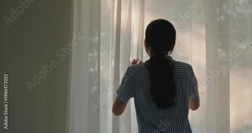 cheerful young Asian woman standing by the window and happily opening both curtains to let the soft natural morning light into her house. 