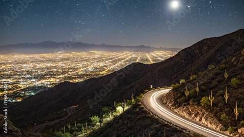 Winding mountain road under a starry night sky overlooks a sprawling lit city. and distant hill glow