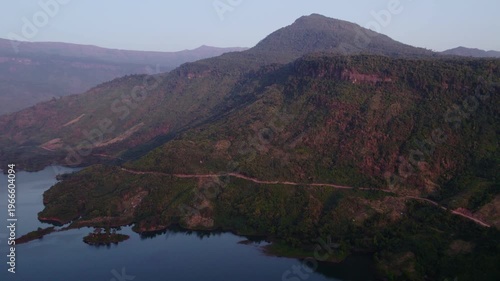 Very high altitude aerial view of dirt roand running along the coast line of large lake with tropical landcsape in the surrounding.
