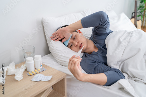 Asian young man lying down on bed in bedroom, looking sick and unwell. 