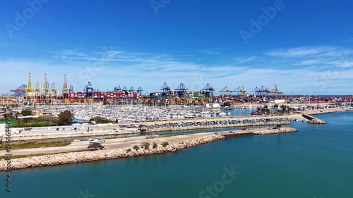 Elevated view of Valencia's large container terminal with colorful stacked cargo and cranes beside a sailboat marina on the Mediterranean coast