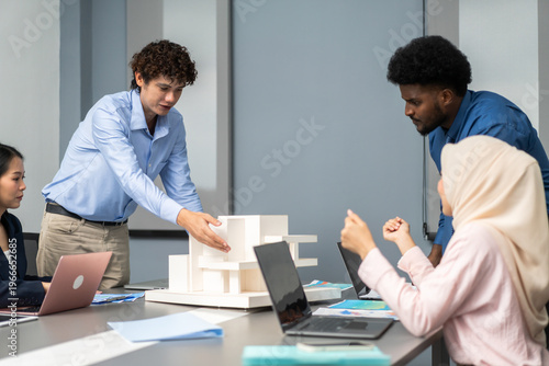 Young male architect presenting white building scale model to diverse business team in office meeting, Corporate teamwork, professional project pitching and real estate development business concept