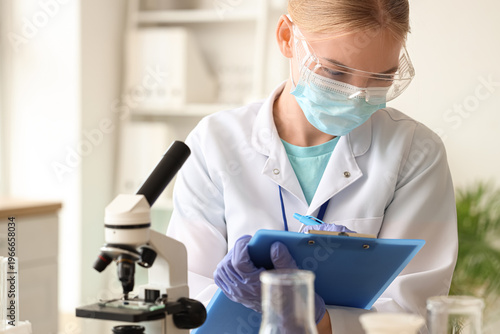 Female scientist with medical mask and microscope writing on clipboard in lab...