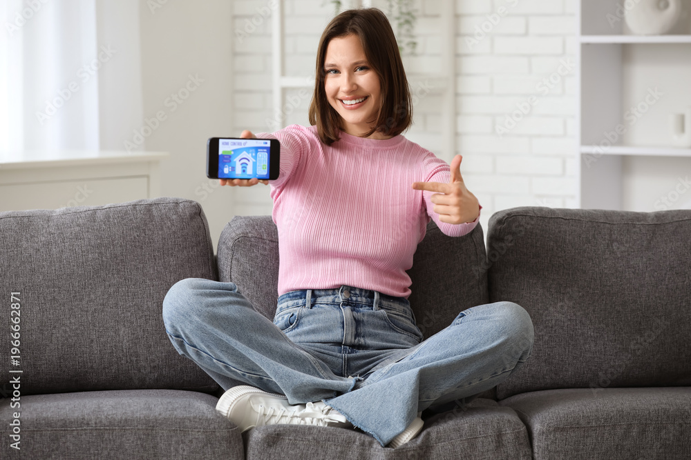 Obraz premium Young happy woman pointing at mobile phone with smart home security system control panel on sofa in room