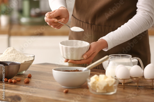 Woman adding sugar into bowl for preparing chocolate brownie at table in kitchen. Closeup