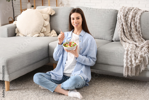 Young woman with bowl of vegetable salad and fork sitting on floor in living ...
